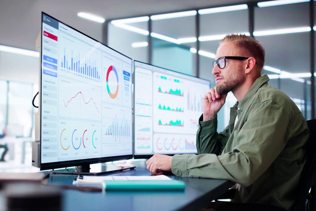 A man with glasses sits at a desk, analyzing data on dual monitors displaying graphs and charts in an office setting.